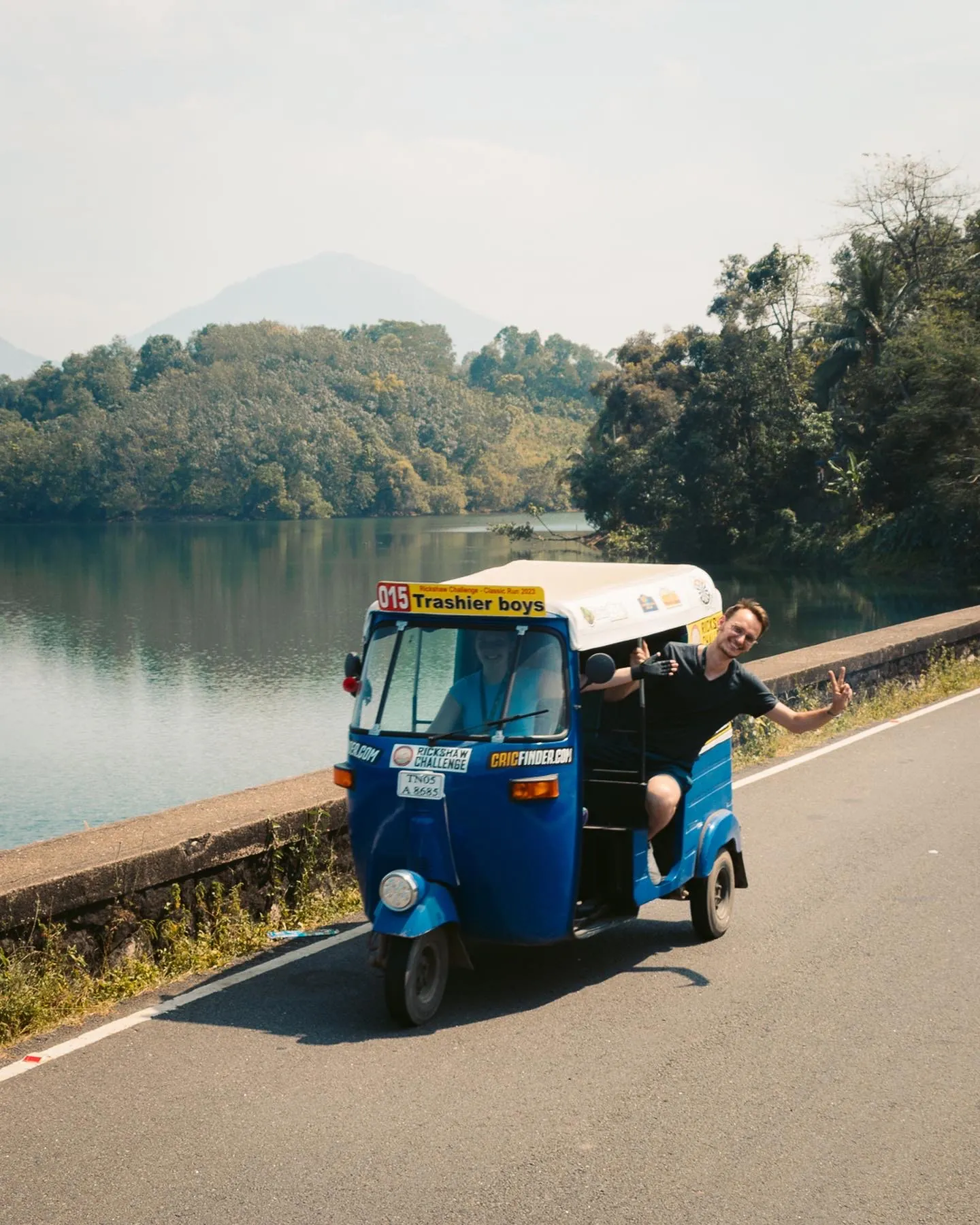 Marius waving from a blue tuk-tuk on a lakeside road