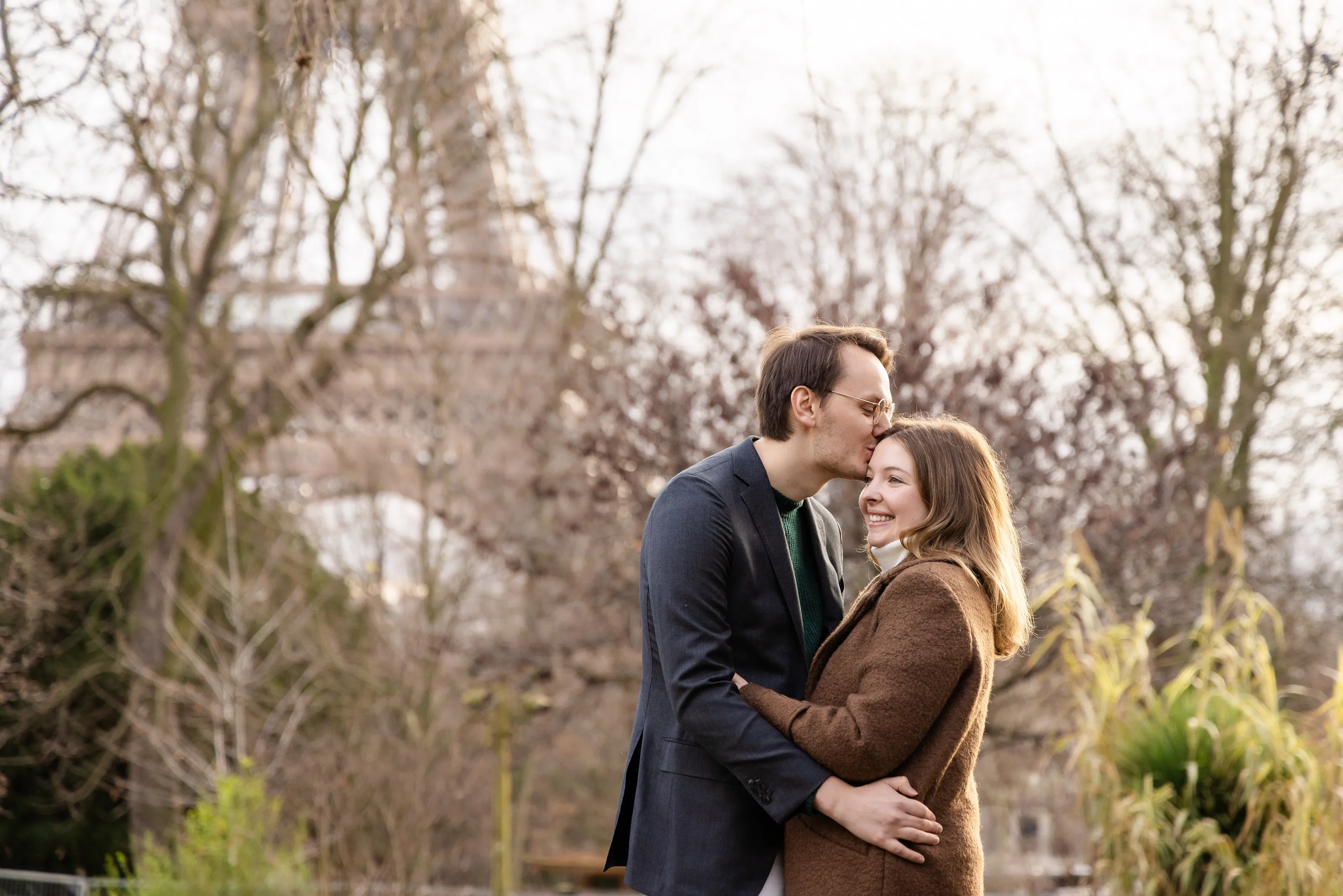 Marius kissing Ally on the forehead in a Paris park, the Eiffel Tower behind them